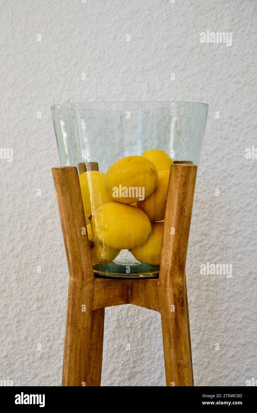 Bol en verre décoratif ou vase avec pieds en bois remplis de citrons frais sur fond de mur blanc. Banque D'Images