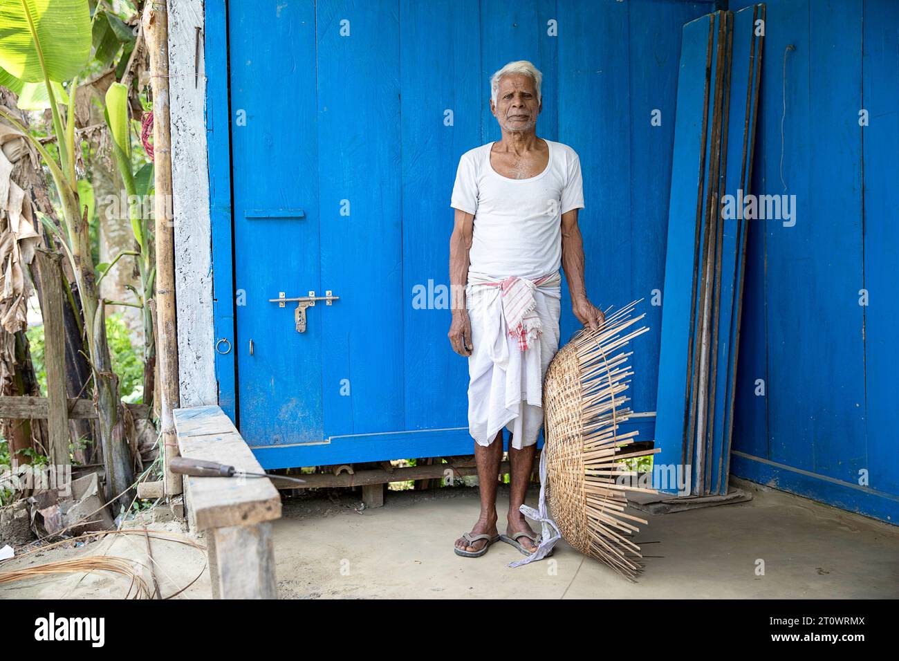 Homme local devant un mur en bois de couleur bleue avec un panier tissé à la main dans ses mains, île Majuli, rivière Brahmapoutre, Assam, Inde Banque D'Images