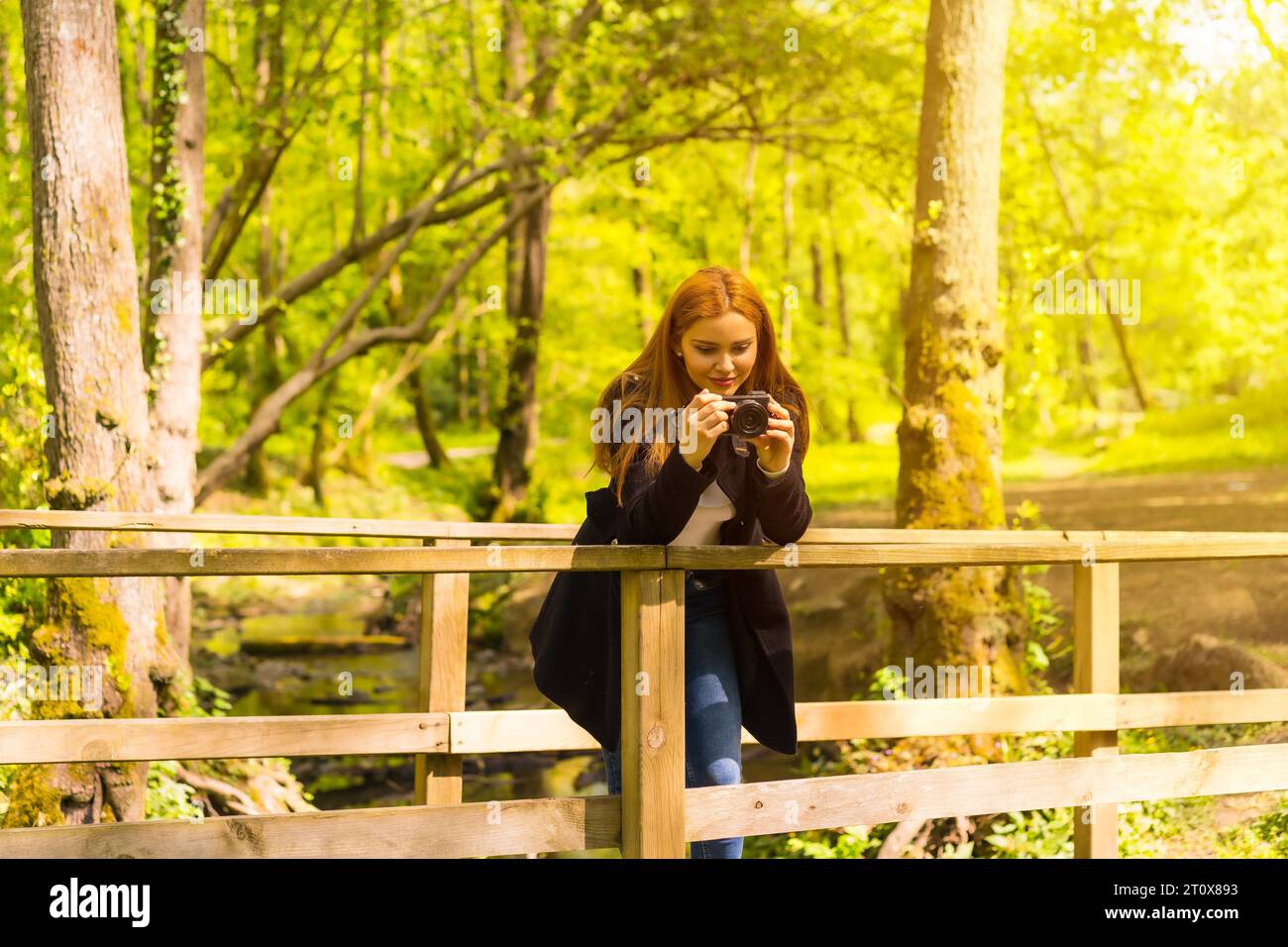 Femme photographe avec une veste noire profitant dans un parc d'automne, prenant des photos sur un pont en bois Banque D'Images