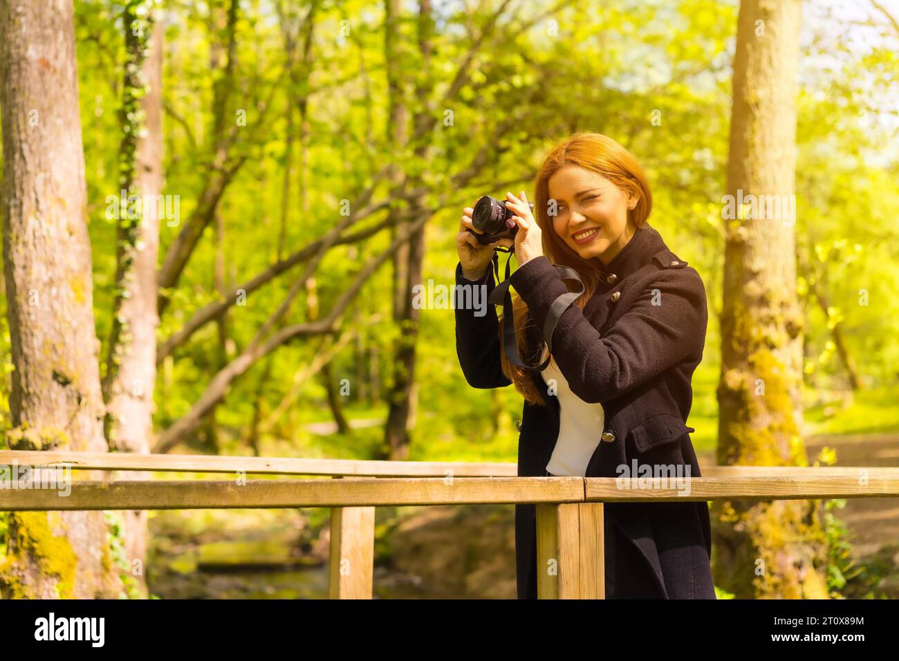 Femme photographe avec une veste noire profitant dans un parc d'automne, prenant des photos au coucher du soleil sur un pont en bois Banque D'Images