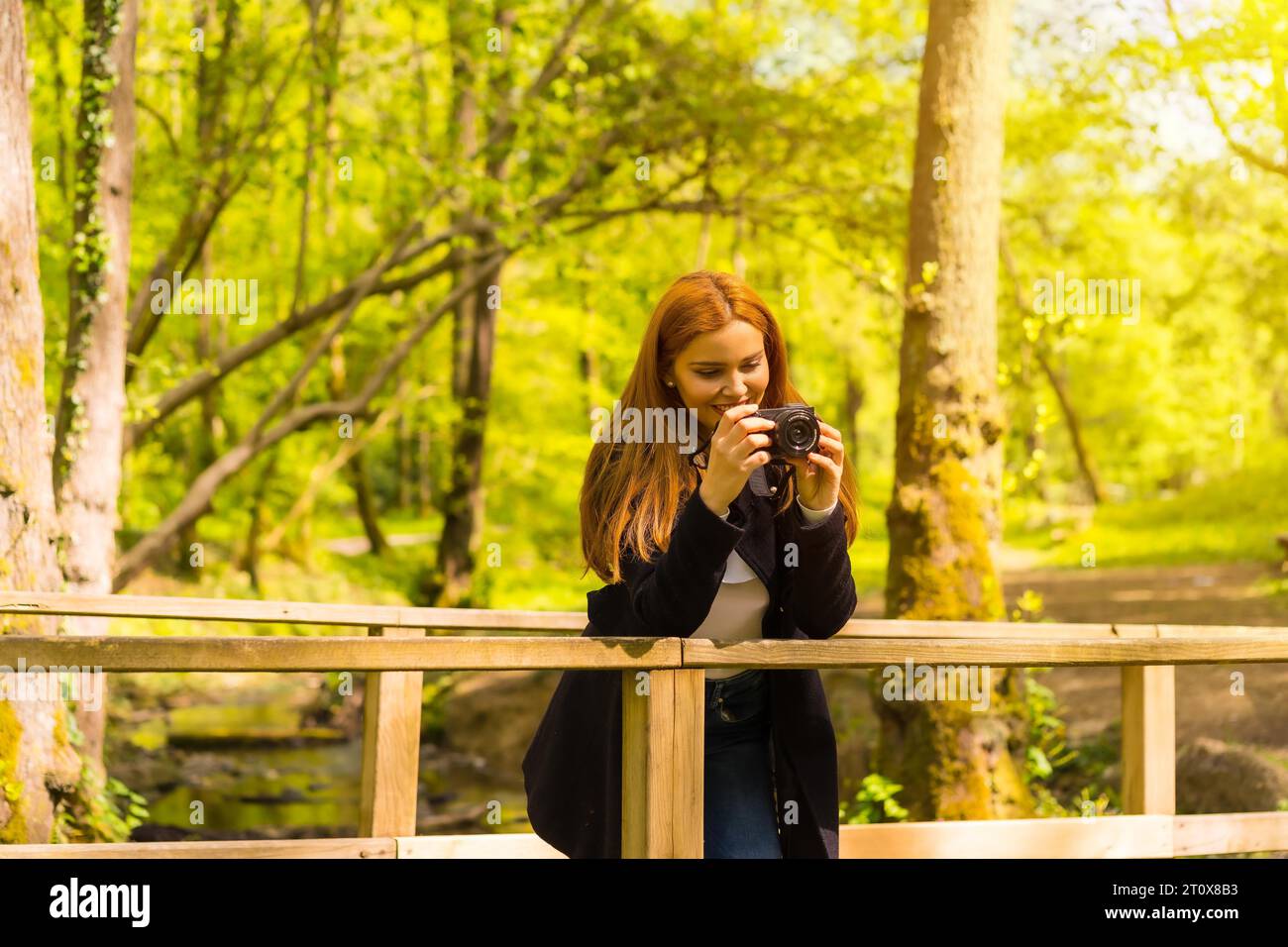 Femme photographe avec une veste noire profitant dans un parc d'automne, prenant des photos sur un pont en bois Banque D'Images