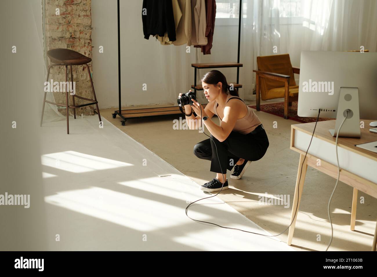 Jeune femme brune avec photo-caméra prenant des photos du modèle tout en étant assis sur des squats dans le centre du studio spacieux pendant la session de photo Banque D'Images