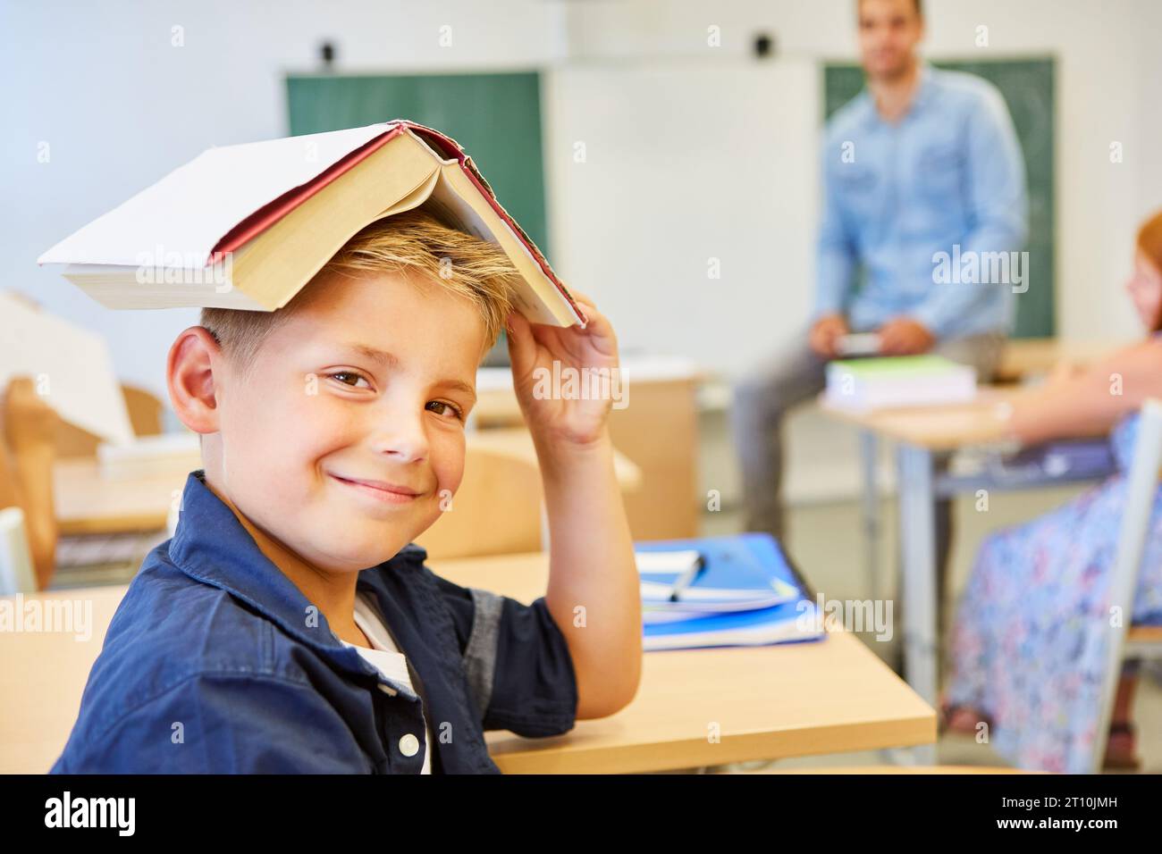 Un garçon souriant drôle avec un livre sur la tête dans une classe d'école primaire comme clown de classe Banque D'Images