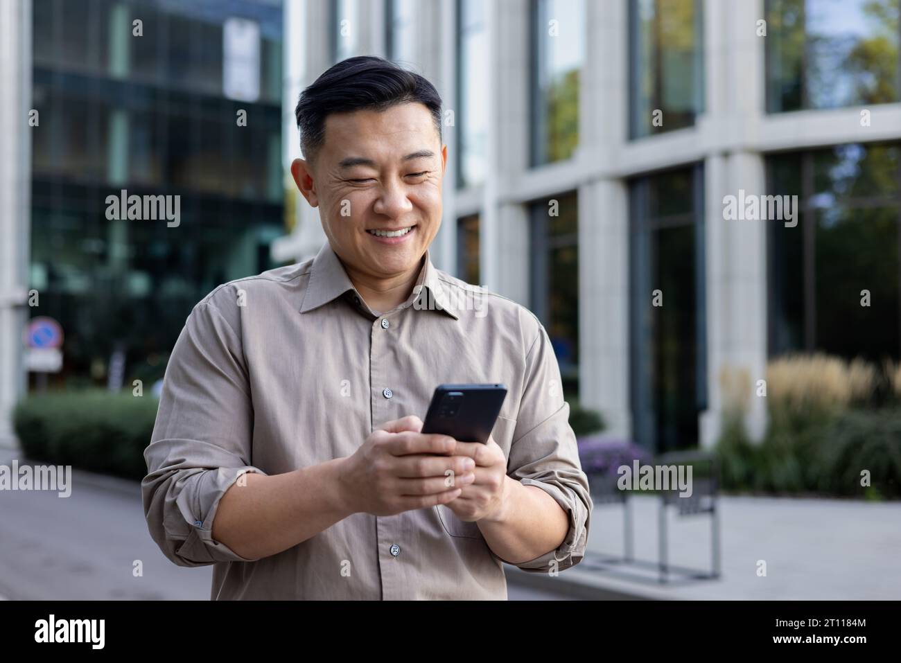 Un jeune asiatique souriant tient un téléphone portable dans ses mains et tape des messages, des SMS, des appels. Debout souriant dans la rue de la ville. Banque D'Images
