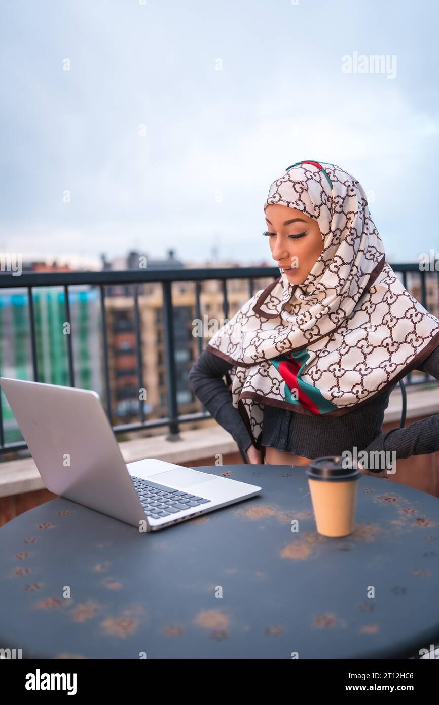 Fille arabe dans un voile blanc à l'ordinateur sur la terrasse d'un magasin de café, faisant un appel vidéo. Jeune arabe moderne avec les nouvelles technologies, photo verticale Banque D'Images