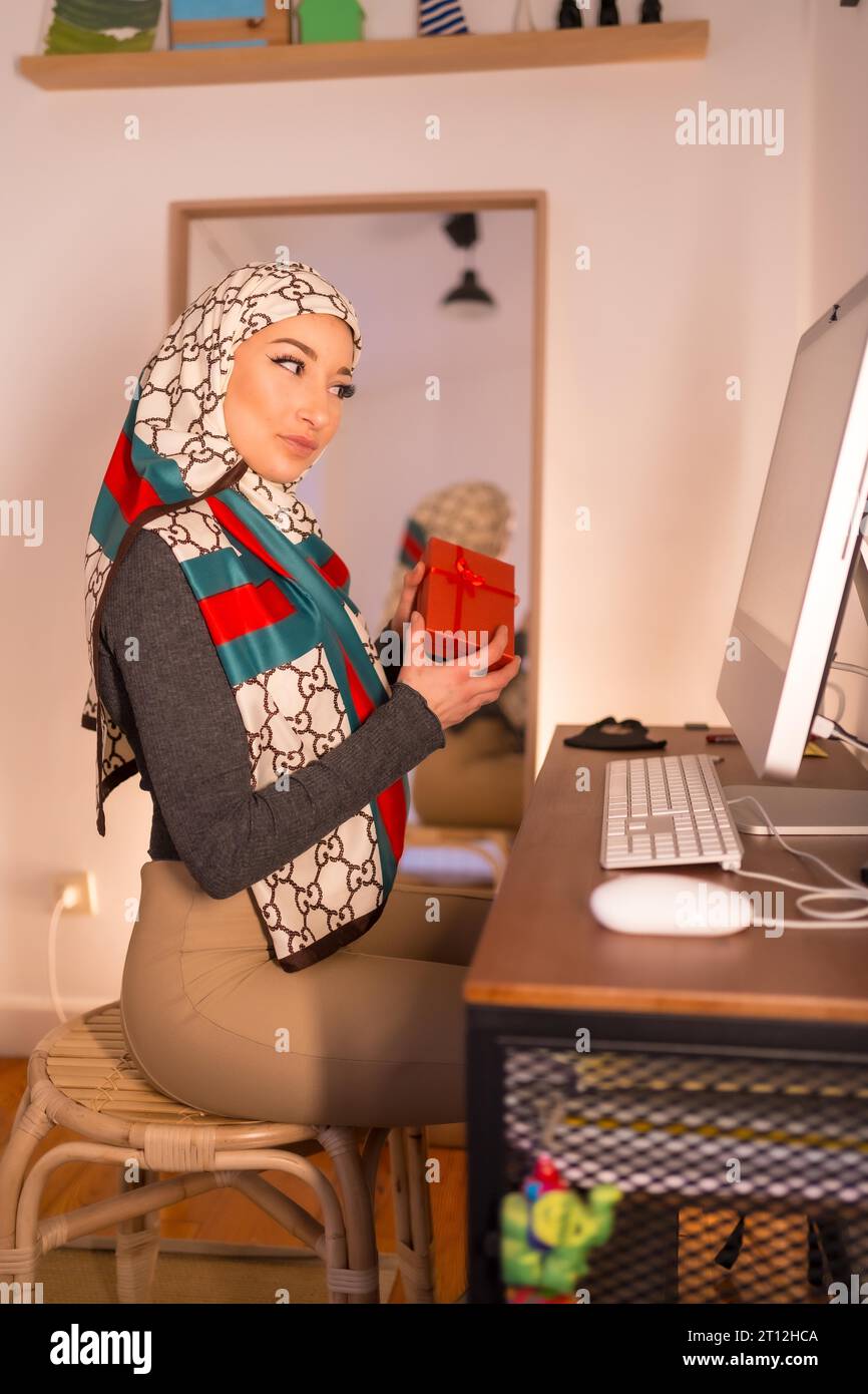 Fille arabe avec un voile blanc à son ordinateur à la maison, faisant un appel vidéo. Jeune arabe moderne avec de nouvelles technologies, ouvrant un cadeau Banque D'Images