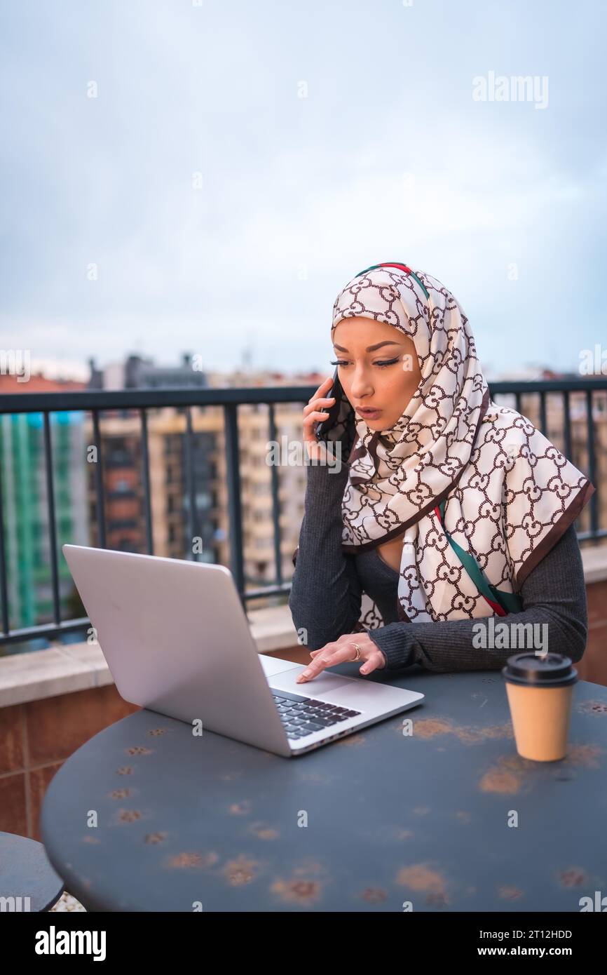 Fille arabe dans un voile blanc à l'ordinateur sur la terrasse d'un magasin de café, faisant un appel vidéo. Jeune arabe moderne avec les nouvelles technologies, photo verticale Banque D'Images