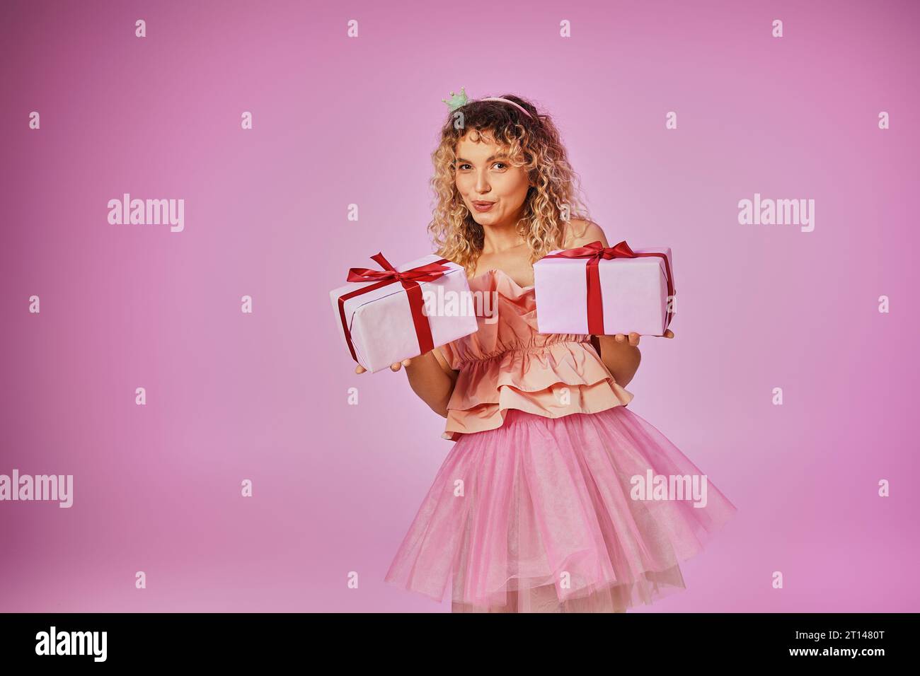 femme blonde rêveuse en tenue rose de fée de dent montrant deux cadeaux sur la caméra sur fond rose Banque D'Images