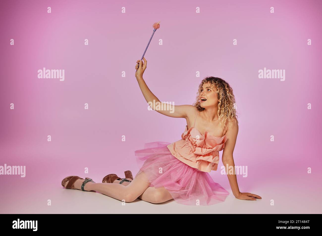 blonde joyeuse avec les cheveux bouclés posant sur fond rose en costume de fée de dent avec baguette magique Banque D'Images