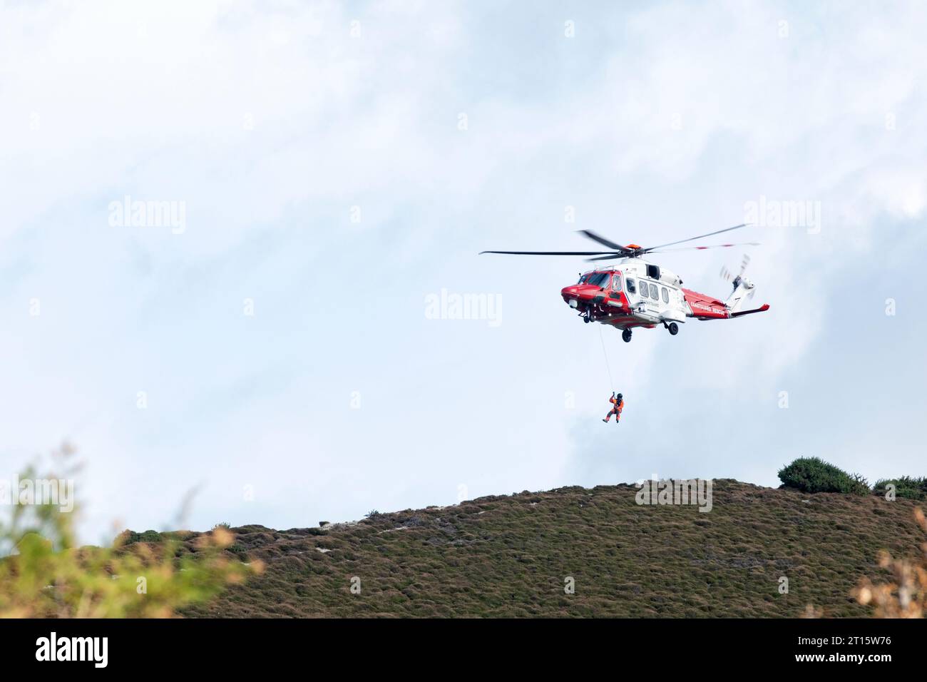 Un hélicoptère HM Coastguard rouge et blanc planant au-dessus d'un promontoire. L'hélicoptère ramène un membre d'équipage à l'aéronef à partir du sol Banque D'Images