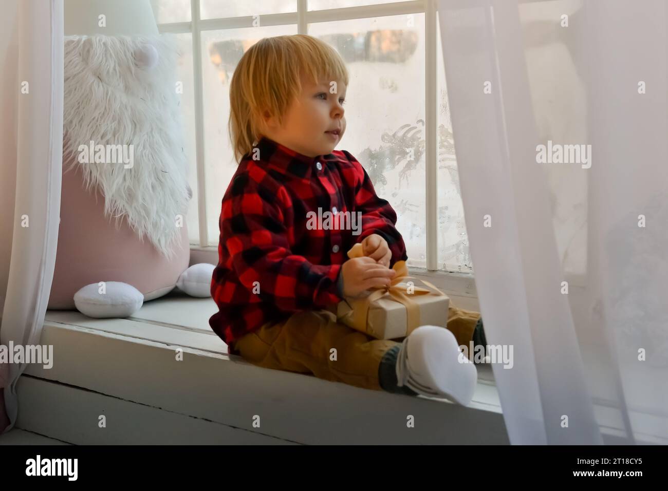 L'enfant est assis sur le rebord de la fenêtre avec un gnome de Noël et tient un cadeau de Noël dans ses mains. Concept de Noël familial Banque D'Images