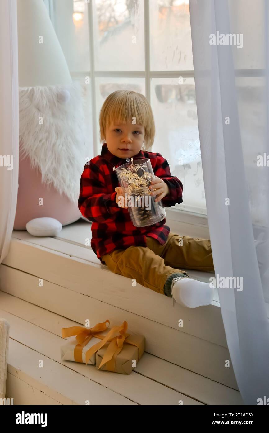 L'enfant est assis sur le rebord de la fenêtre avec un gnome de Noël et tient une lanterne de Noël et un cadeau du nouvel an, boîte dans ses mains. Concept de Noël familial Banque D'Images