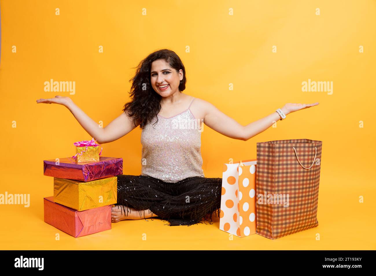 Portrait d'une femme indienne asiatique excitée assise sur le sol avec des sacs à provisions et une boîte cadeau isolée sur fond orange, concept Shopper ou shopaholic Banque D'Images