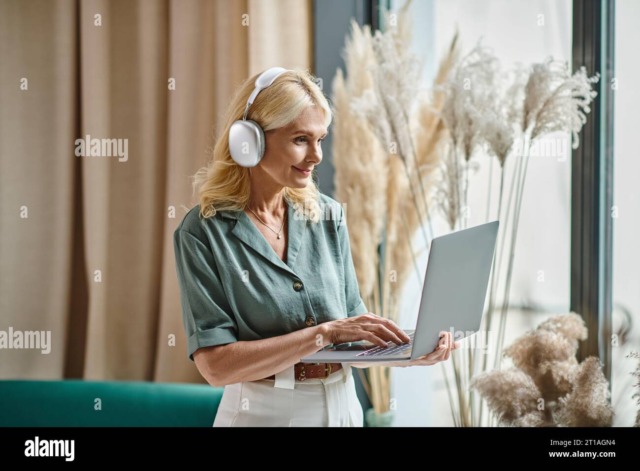 joyeuse femme d'âge moyen dans un casque sans fil écoutant de la musique et utilisant un ordinateur portable à la maison Banque D'Images