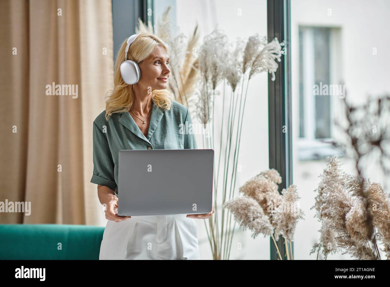 heureuse femme d'âge moyen dans un casque sans fil écoutant de la musique et tenant l'ordinateur portable à la maison Banque D'Images