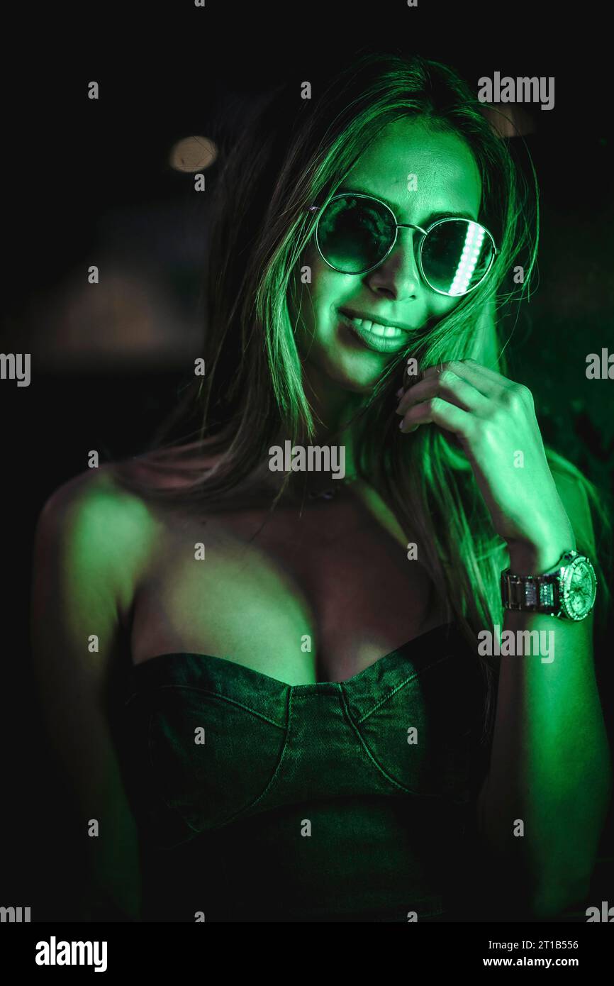 Brunette caucasienne dans une robe noire éclairée par une lumière LED verte réfléchie par des cristaux noirs. Photographie nocturne urbaine. Fille avec lunettes de soleil et Banque D'Images