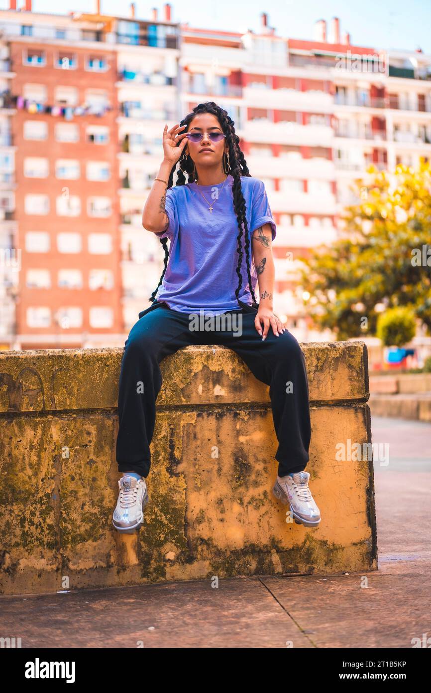 Jeune homme à la peau foncée dans un T-shirt violet avec de longues tresses et des lunettes violettes assis sur un loft en ciment jaune Banque D'Images