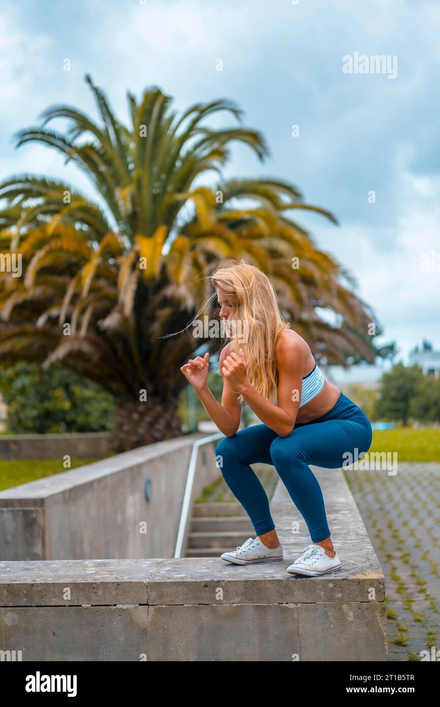 Séance de fitness avec une jeune femme blonde caucasienne s’exerçant dans un parc avec des palmiers avec un maya bleu sur les pieds et une chemise courte blanche. En cours Banque D'Images