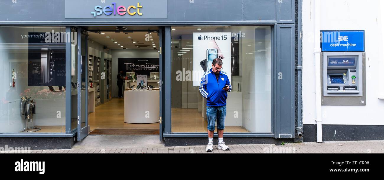 Une image panoramique d'un homme debout devant un magasin de téléphonie mobile regardant son téléphone dans une rue du centre-ville de Truro à Cornwall au Royaume-Uni. Banque D'Images