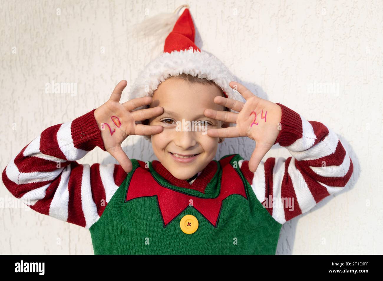 Enfant mignon souriant dans le chapeau de Père Noël et pull elfe, les numéros de la nouvelle année 2024 sont écrits sur les paumes Banque D'Images