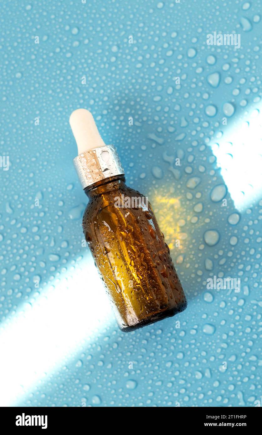 Bouteille de sérum, huile cosmétique sur fond bleu avec lumière dure et gouttes d'eau. avec ombres à plat, vue de dessus Banque D'Images