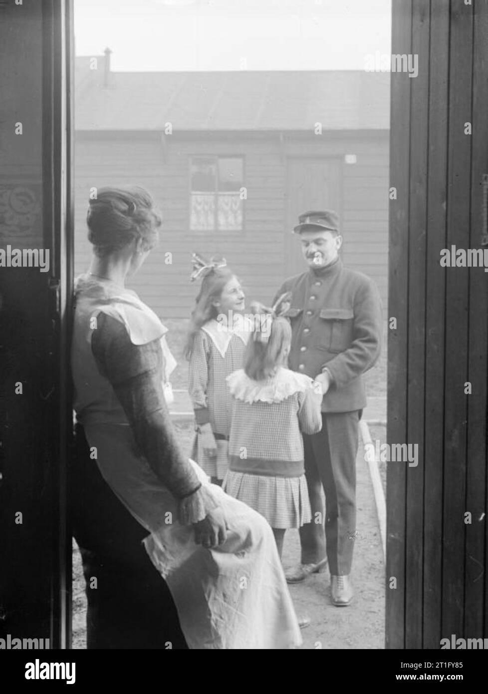 Les travailleurs civils belges en Grande-Bretagne durant la Première Guerre mondiale, deux jeunes filles belges saluer leur père à l'extérieur de la maison à son retour de travail, comme leur mère a l'air sur l'encadrement de la porte. Il s'agit d'un travailleur de munitions et la famille habite à Elisabethville, une zone clôturée de Birtley, près de Gateshead, dans le comté de Durham, composé entièrement de réfugiés belges et des hommes blessés, produisant des coquilles à l'effort de guerre britannique. Cette "colonie" a été nommée d'après la Reine de Belgique et a été exécuté par le gouvernement belge. Dans le fond, le style simple de l'habitation familiale en bois peut être vu. Banque D'Images