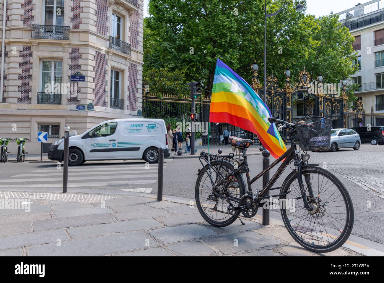 Vélo garé au premier plan, portant un drapeau arc-en-ciel avec le mot 'paix' ('paix'). En arrière-plan, les voitures passent devant l'entrée ornée d'un parc parisien. Banque D'Images