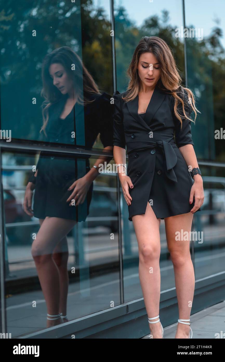Street style, une jeune femme caucasienne entreprenante entrant dans le bureau pour travailler dans un costume noir et des talons. Reflété dans le verre du verre noir Banque D'Images