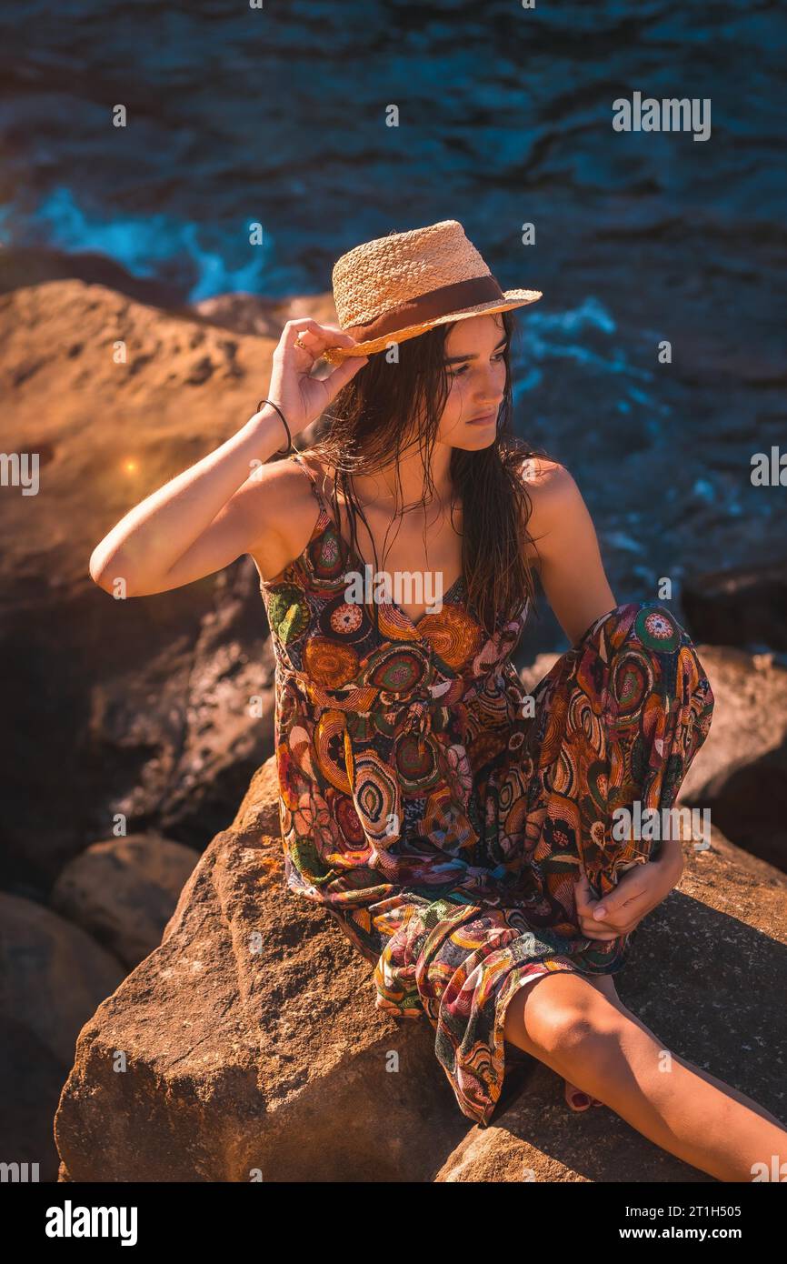 Une jeune femme caucasienne avec les cheveux mouillés, une robe de fleurs et un chapeau assis sur un rocher au bord de la mer, style de vie estival à l'heure d'or Banque D'Images