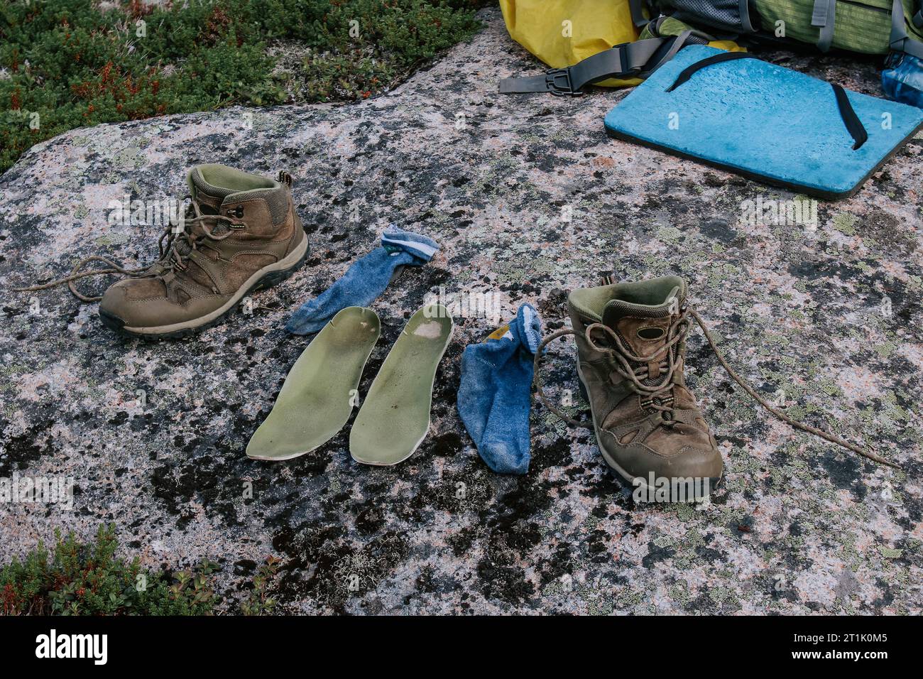 Vieilles bottes de trekking brunes, semelles vertes et chaussettes bleues à sécher après une journée de randonnée, disposées sur pierre. Pieds mouillés en voyage. Camping de voyage Banque D'Images