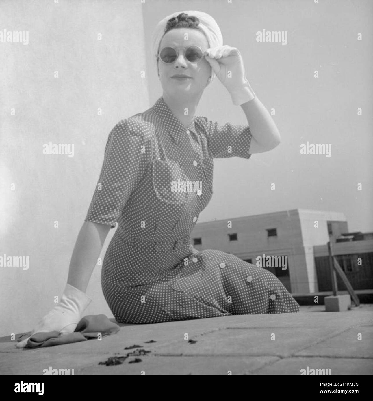 Comment une femme britannique s'habille de Vêtements de guerre- Utilitaire en Grande-Bretagne, 1943, un modèle est assis sur un mur qu'elle pose pour la caméra sur un toit à Bloomsbury, à montrer sa scarlet et point blanc-utilitaire imprimé robe rayonne avec boutonnage. Cette robe coûte 7 coupons et 53/-. Elle porte la robe avec un turban blanc (également en vedette dans D 14782, 14786 et 14798 D D) et gants blancs. Elle est également doté d'une paire de lunettes de soleil, sa main gantée tenant le bras de fil des verres. Banque D'Images