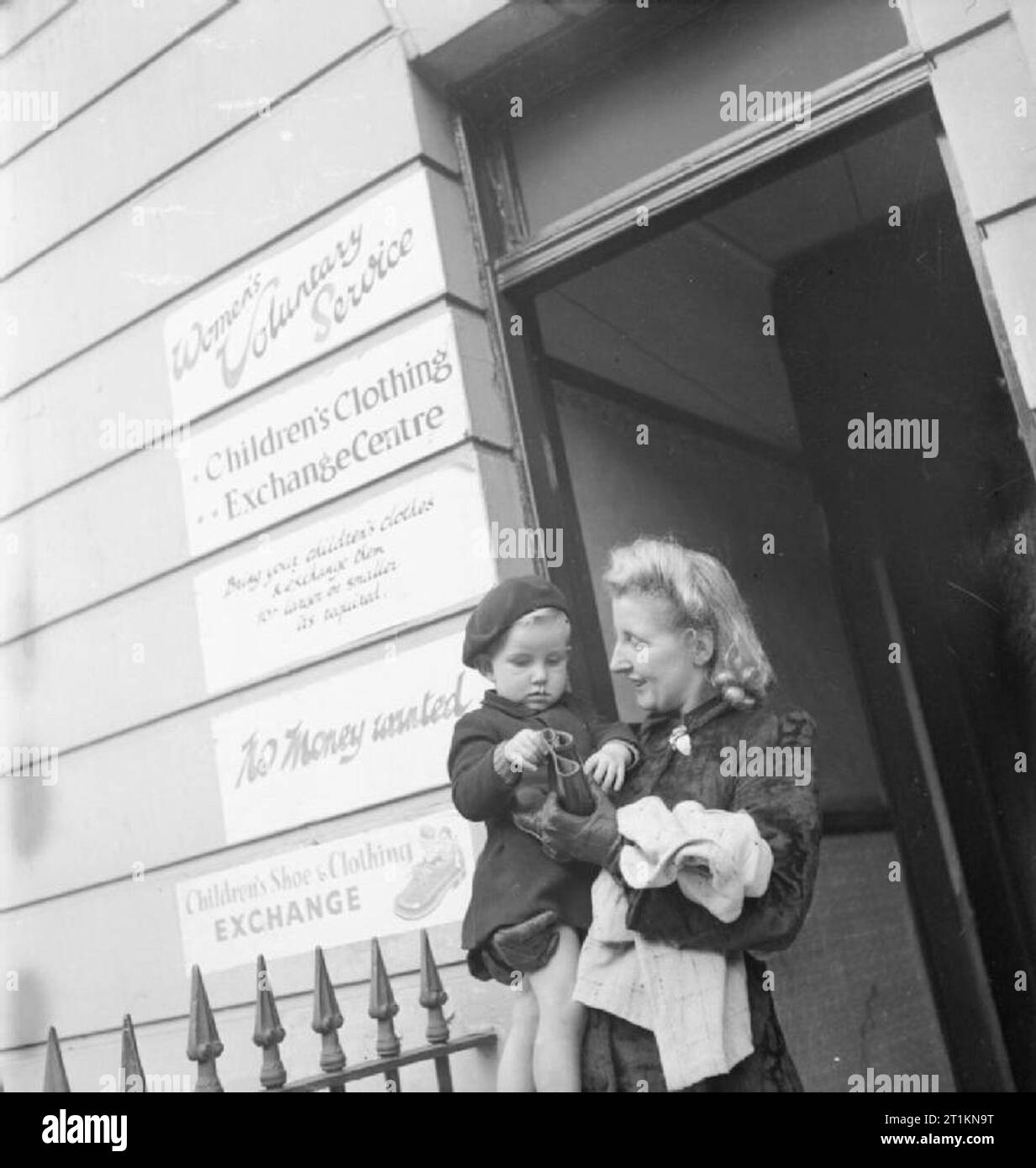 Marketing sans argent- Échange de vêtements pour enfants, Norwood, Angleterre, RU, 1943 Un jeune garçon et sa mère à l'extérieur d'un échange de vêtements pour enfants gérés par la Women's Voluntary Service à Norwood, dans le sud de Londres. Ils ont échangé un tas de bébé woollies et certains de ses hors-cultivé pour un manteau épais et une paire de bottes. Banque D'Images