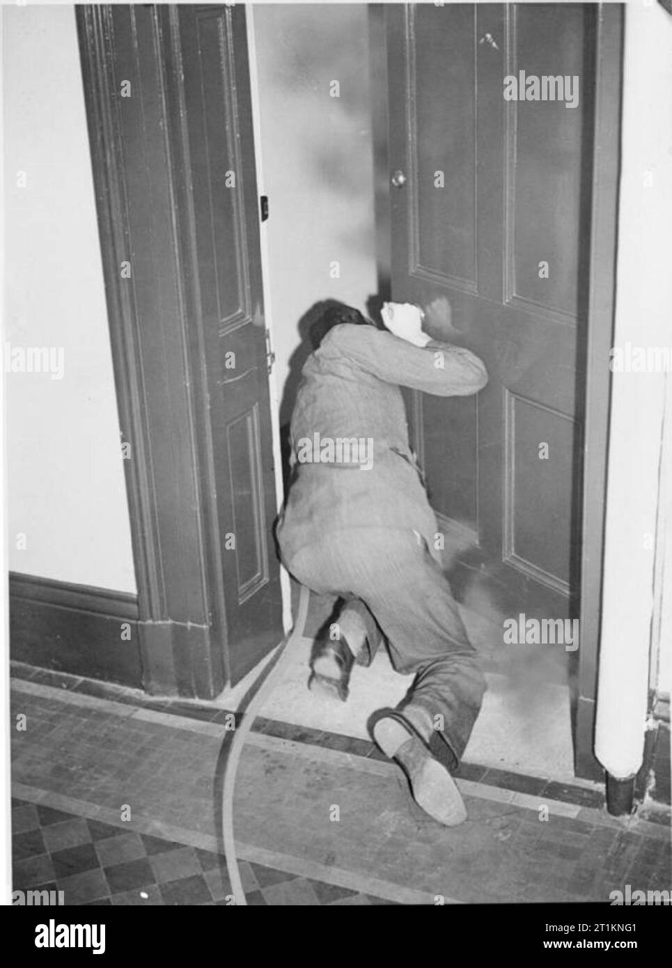 Précautions de protection de l'air et de la défense civile des guignols de l'avant Accueil- Comment éteindre un feu, bombe C 1940 Un homme s'accroupit devant la porte d'une pièce remplie de fumée et vise sa pompe l'étrier autour du bord de la porte pour tenter d'éteindre le feu. Banque D'Images