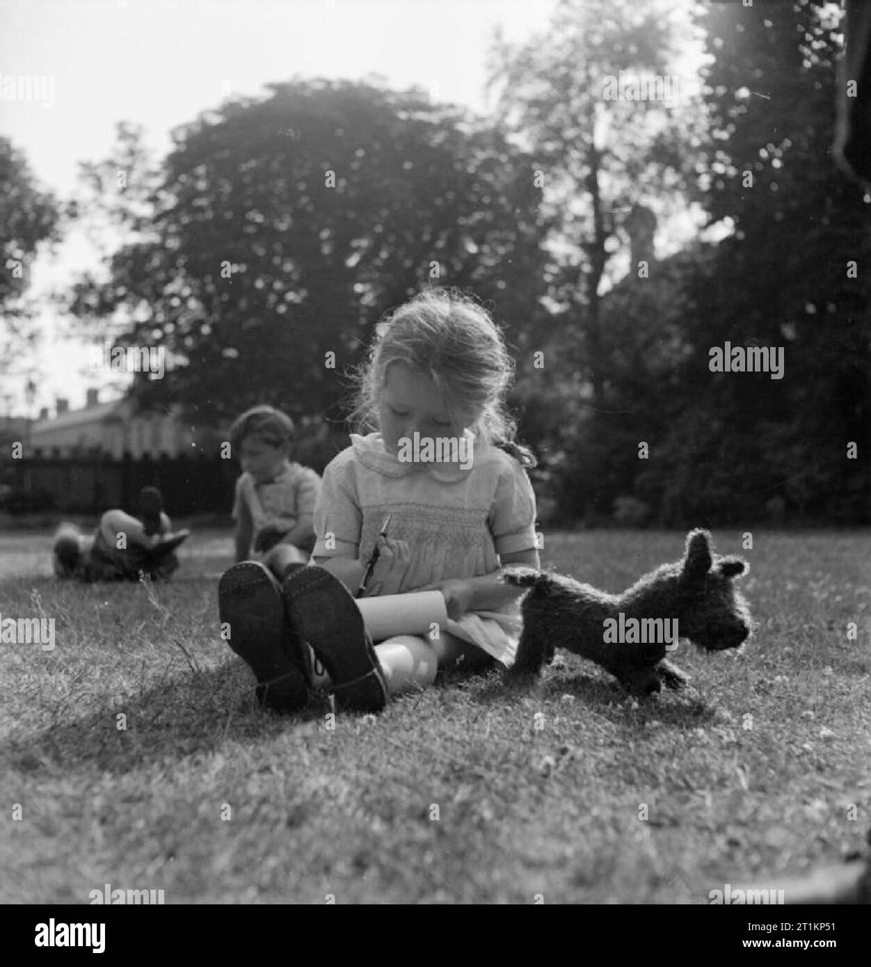 Garderie Pour les mères au travail- le travail du silex Green Road, pépinière, Birmingham, 1942 Sylvia Rainford (de 4) est assis sur l'herbe dans la soirée le soleil, son jouet chien à ses côtés, et écrit une lettre à son père, qui est au service de quelque part au Moyen-Orient. Sylvia vit à la pépinière pendant que sa mère ne le travail de nuit dans une fabrique de munitions. Banque D'Images