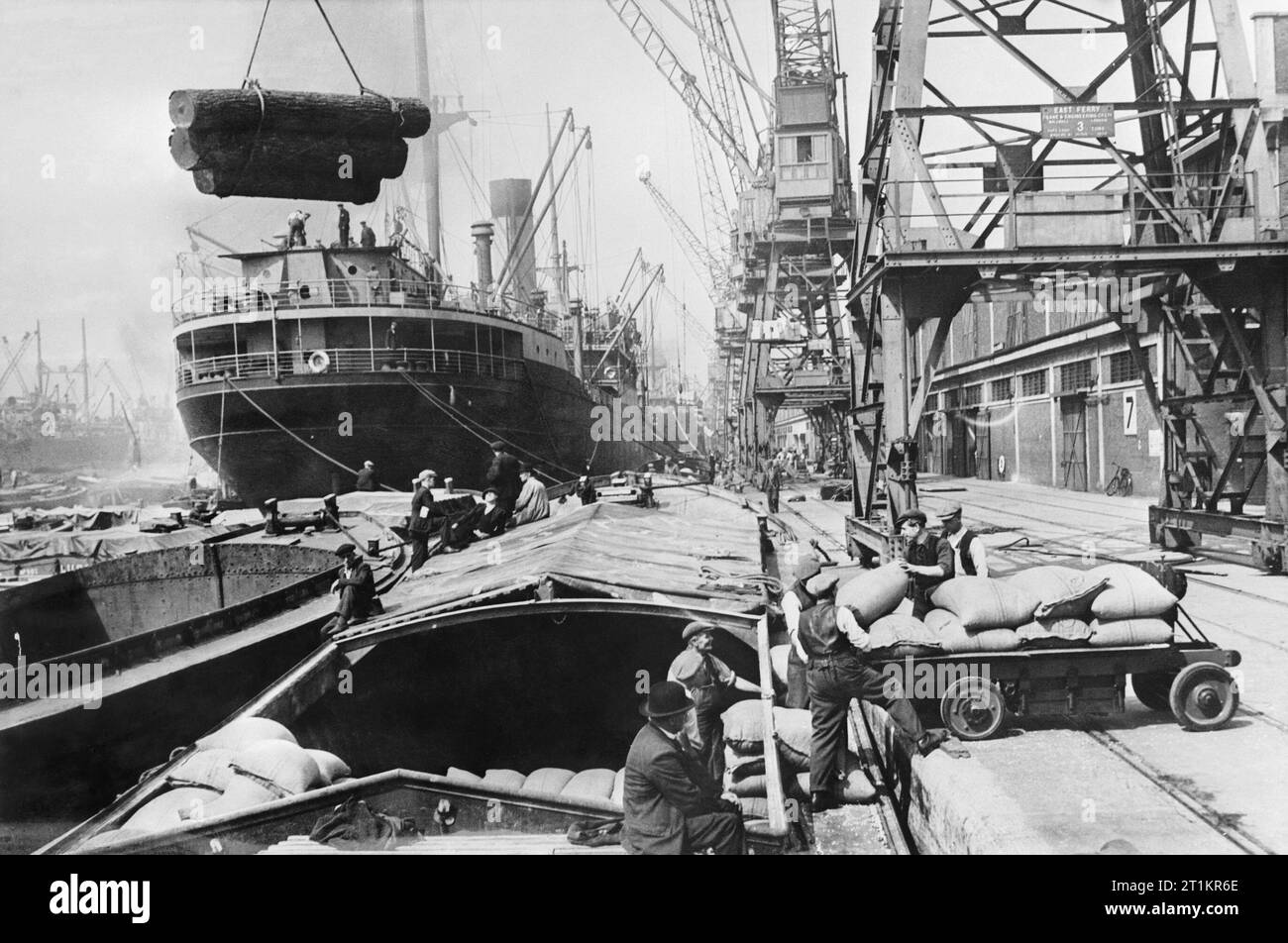 Scène dans un chantier naval britannique pendant la Seconde Guerre mondiale, les quais transfèrent à la main, au véhicule et à la grue des marchandises importées et exportées entre des navires marchands et des barges fluviales à un chantier naval non identifié, éventuellement sur la côte ouest. Commentaire : voir le libellé sur la grue. 'East Ferry'. Il peut s'agir de Millwall ou de Newport Docks. Voir https://www.gracesguide.co.uk/East Ferry Road Engineering Works Co Banque D'Images