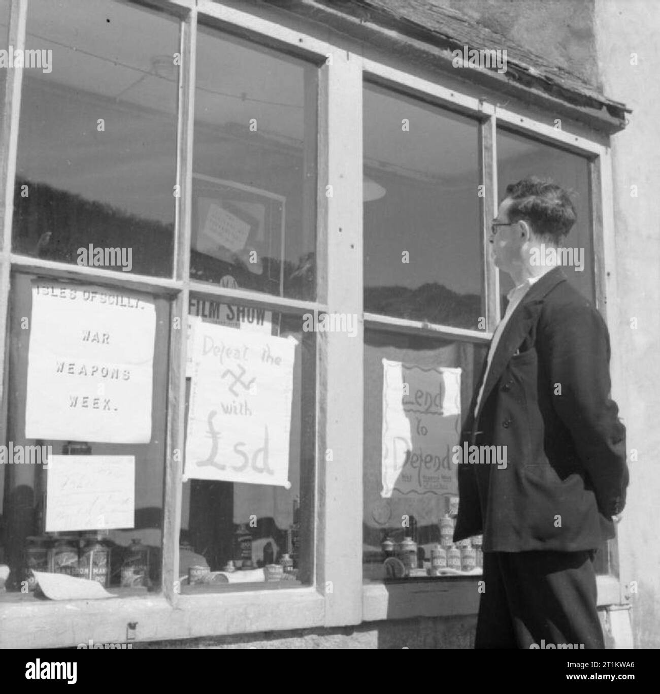 Wartime in the Scillies- la vie quotidienne sur les îles Scilly, avril 1941 Un Scillonian regarde des affiches faites maison pour la semaine des armes de guerre, qui sont exposées dans une vitrine sur Tresco, Scilly Isles, 1941. Les slogans incluent 'prêter à défendre' et 'vaincre le (svastika dessiné ici) avec ?GBPsd'. Banque D'Images