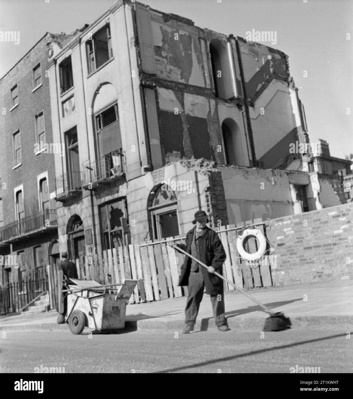 Le nettoyage des rues de la femme- Brigade ramasseurs au travail féminin, Londres, 1942, Mme G Warren De La Rue des Femmes de St Pancras Brigade de nettoyage utilise un grand balai pour balayer la route. Elle est le brossage dans la gouttière à côté de la bordure, avec son dustcart à proximité. Les bâtiments derrière elle montrent des signes d'être durement touchée par des raids aériens. Selon la légende originale, Mme Warren a 35 ans et "charred' dans les bureaux avant la guerre. La photographie a été prise après une pause pour le petit déjeuner entre 8 et 8 h 30. Banque D'Images
