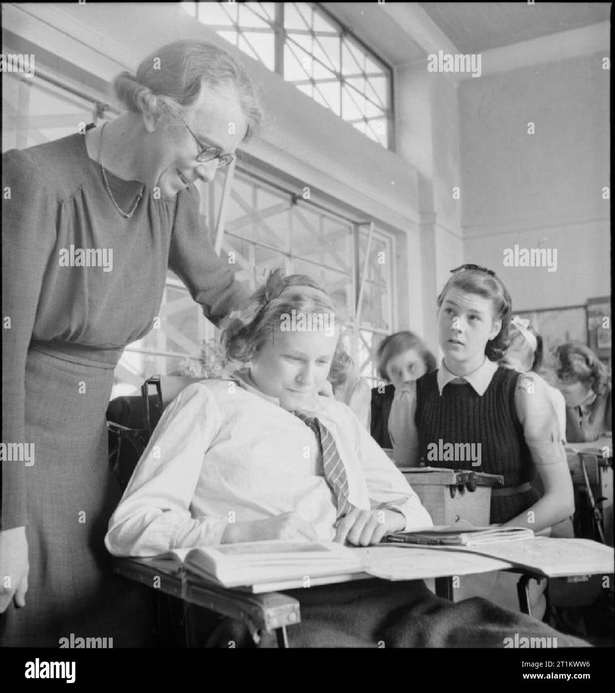 Young Britons Study American History- Education in Wartime England, 1943 Professeur d'histoire Miss L M Molineux aide Monica Young avec son travail lors d'une leçon sur l'histoire américaine à Albany Senior School à Enfield. Monica est handicapée, et a un bureau équilibré sur les bras de son fauteuil roulant, sur lequel écrire. Les panneaux de verre qui séparent la salle de classe du couloir ont des croix de ruban adhésif entrecroisées pour éviter les dommages causés par le souffle. Banque D'Images