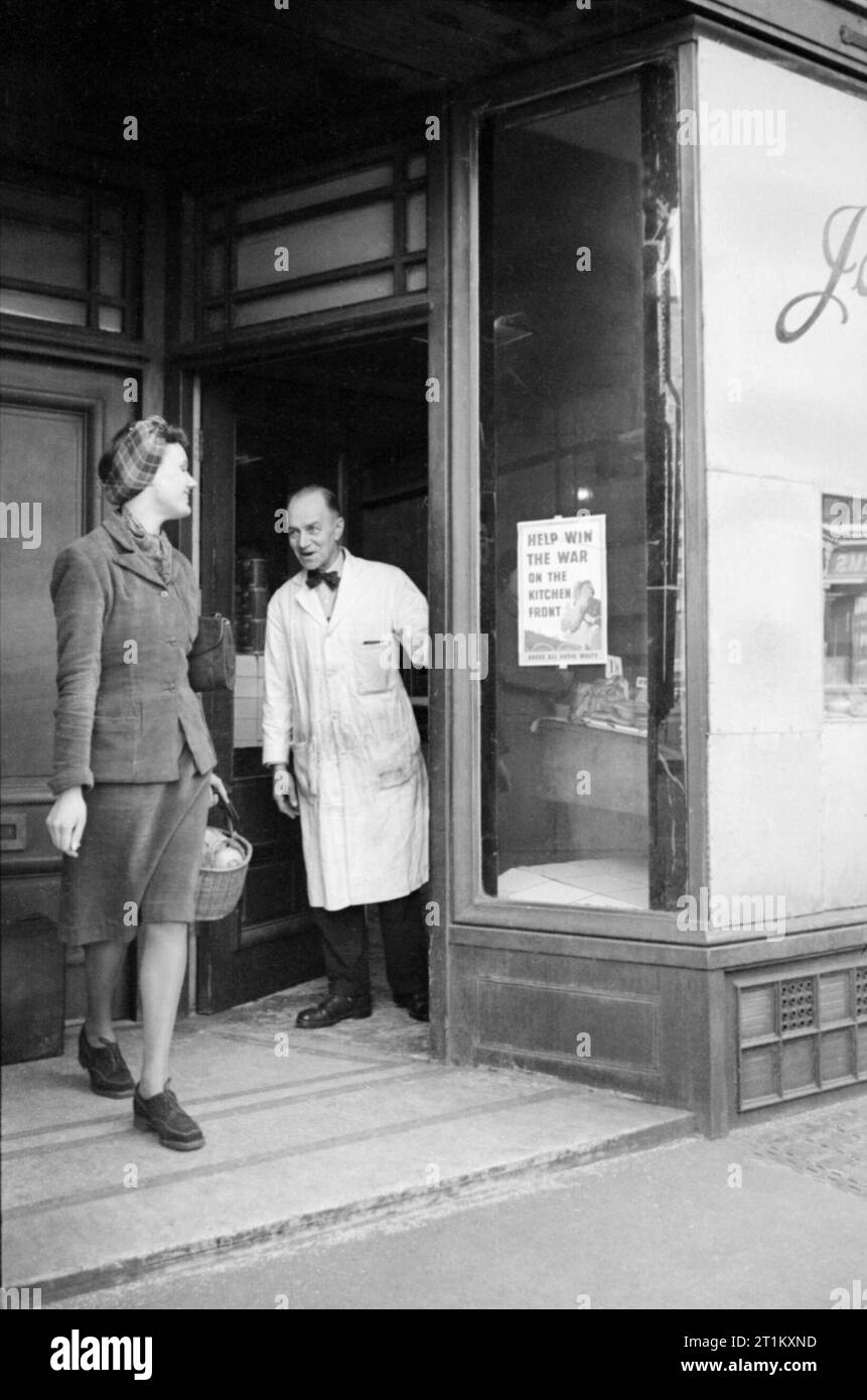 Une femme au foyer de Londres quitte sa boucherie locale après avoir acheté sa ration de viande, février 1941. Mme Day l'a dit au revoir à la boucherie alors qu'elle quitte sa boutique, après avoir acheté sa ration de viande. La fenêtre de la boucherie a été arraisonné plus d'après le verre a été éliminé au cours d'un raid aérien. Le signe dans la fenêtre du boucher dit "aider à gagner la guerre sur la cuisine avant. Par-dessus tout éviter le gaspillage". Cette photographie a probablement été prise sur la King's Road à Chelsea. Banque D'Images