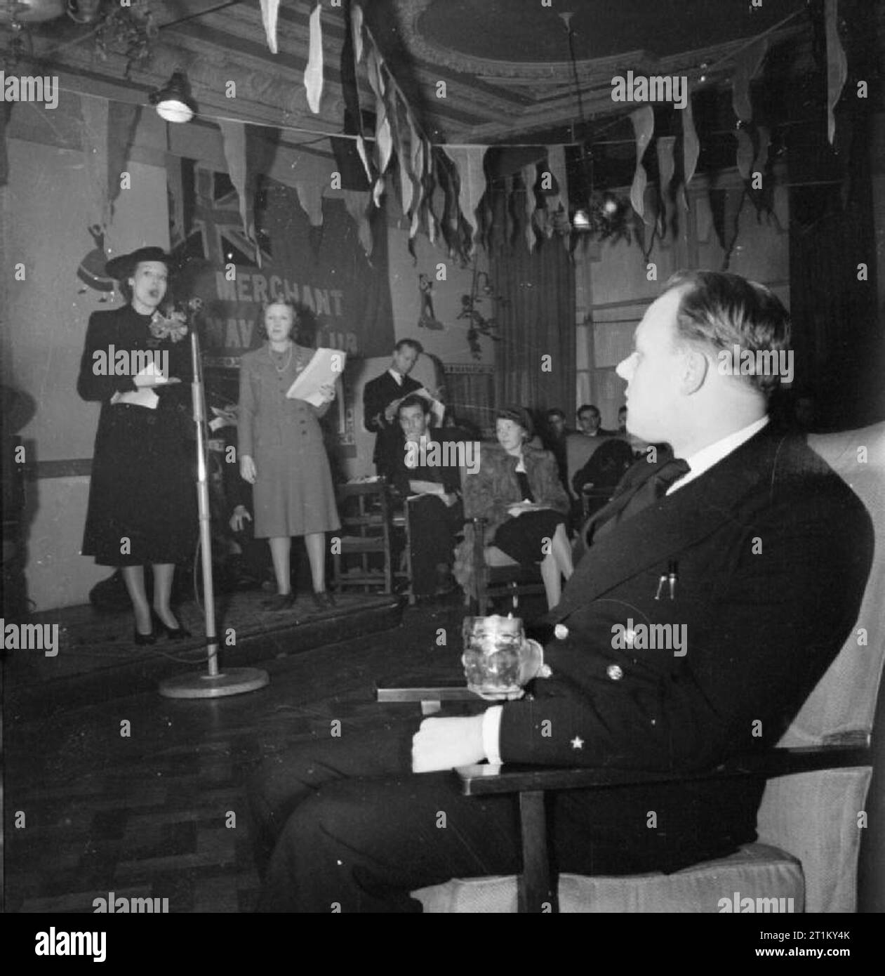 Un marin marchand vient à la ville- le travail de la marine marchande Club, Piccadilly, Londres, 1942 Richard pâtés, 25 ans, marin marchand bénéficie d'un verre de bière tout en écoutant chanter à la Dixon Adele Merchant Navy Club à Piccadilly. Derrière Adele, l'hôtesse, Doris Hare, peut être vu, ordre de marche dans la main. La chambre a un plafond orné de couleurs vives et Bunting a été tendu autour de la salle. Plusieurs autres personnes peut être vu à l'écoute de la performance. Banque D'Images