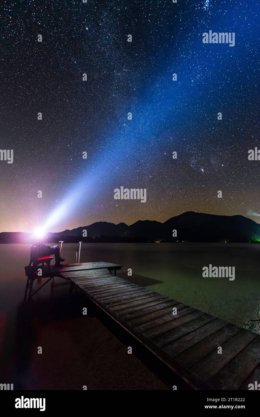 Homme assis sur un banc sur une jetée en bois sur la rive du lac Kochel en Bavière, avec un faisceau lumineux de sa lampe frontale dans le ciel étoilé de nuit Banque D'Images