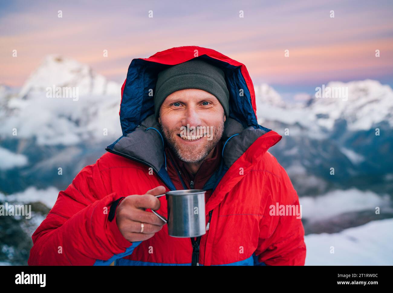 Portrait de souriant à la caméra alpiniste de haute altitude habillé rouge chaude de la veste d'aube tenant tasse en métal de thé chaud dans le backgroun de panorama de montagnes Banque D'Images
