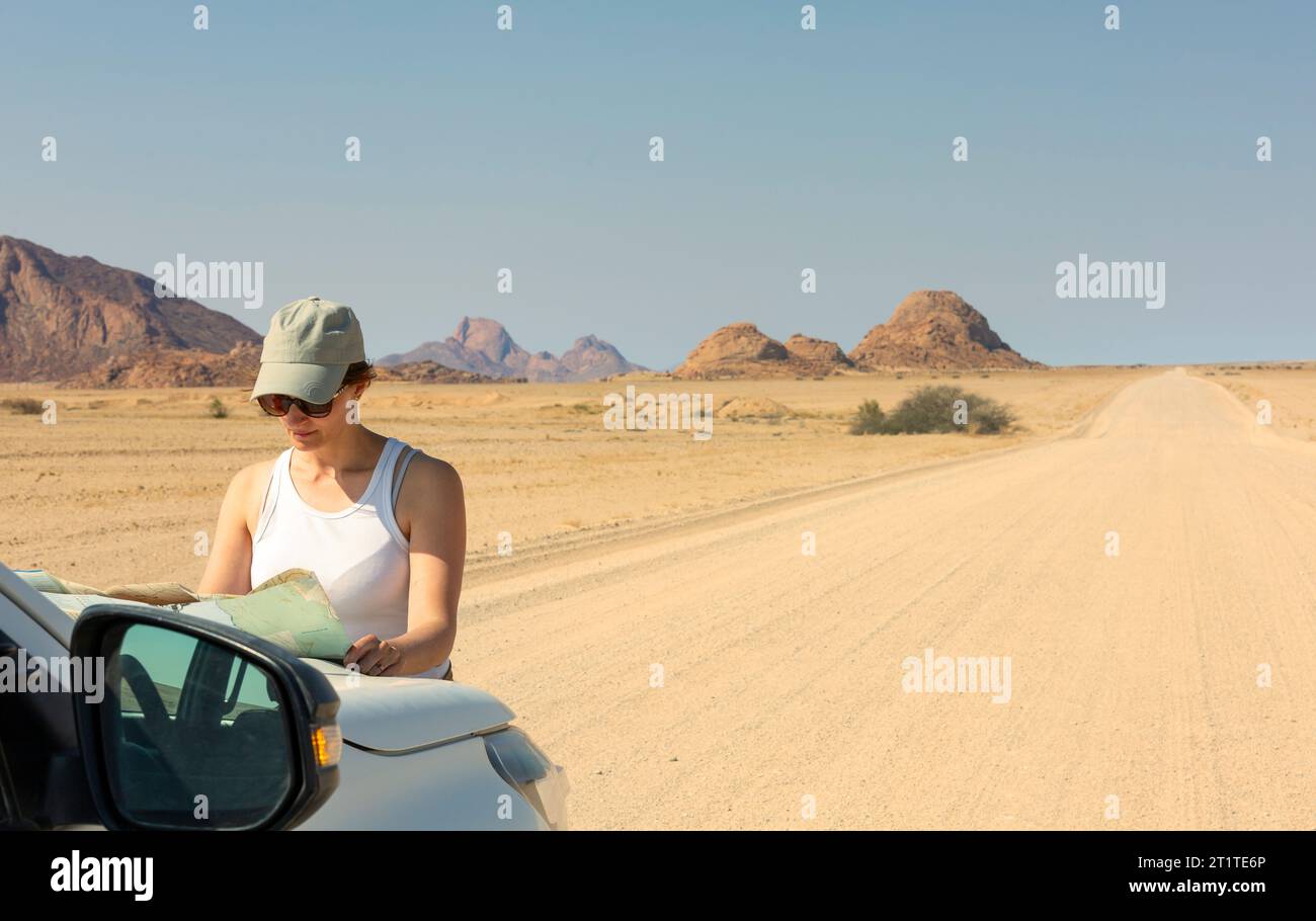 Jeune femme voyageur dans le chapeau regardant dans la carte touristique debout près de la voiture en afrique Banque D'Images Jeune femme voyageur dans le chapeau regardant dans la carte touristique debout près de la voiture en afrique Banque D'Images