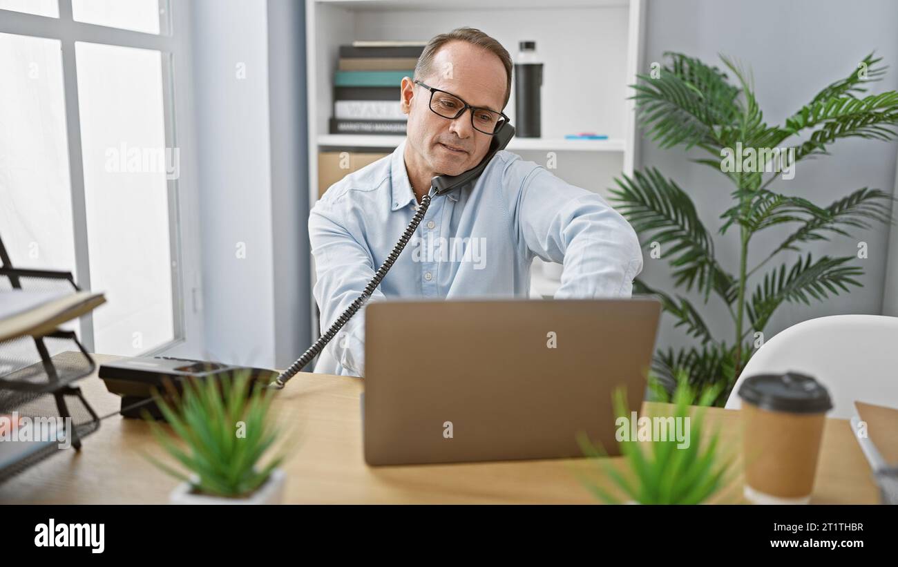 Homme d'affaires d'âge moyen détendu s'engageant dans une conversation téléphonique sérieuse tout en travaillant sur son ordinateur portable au bureau Banque D'Images