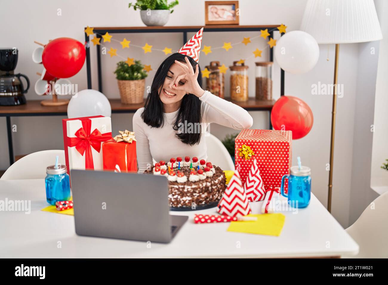 Femme hispanique célébrant l'anniversaire avec un gros gâteau au chocolat faisant appel vidéo souriant heureux faire ok signe avec la main sur l'œil regardant à travers les doigts Banque D'Images