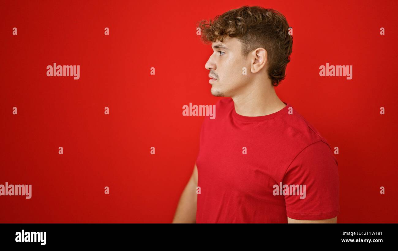 Cool, jeune homme hispanique donnant un regard de côté sérieux debout dans la lumière ensoleillée, dépeignant un style de vie détendu sur un fond de mur rouge isolé Banque D'Images