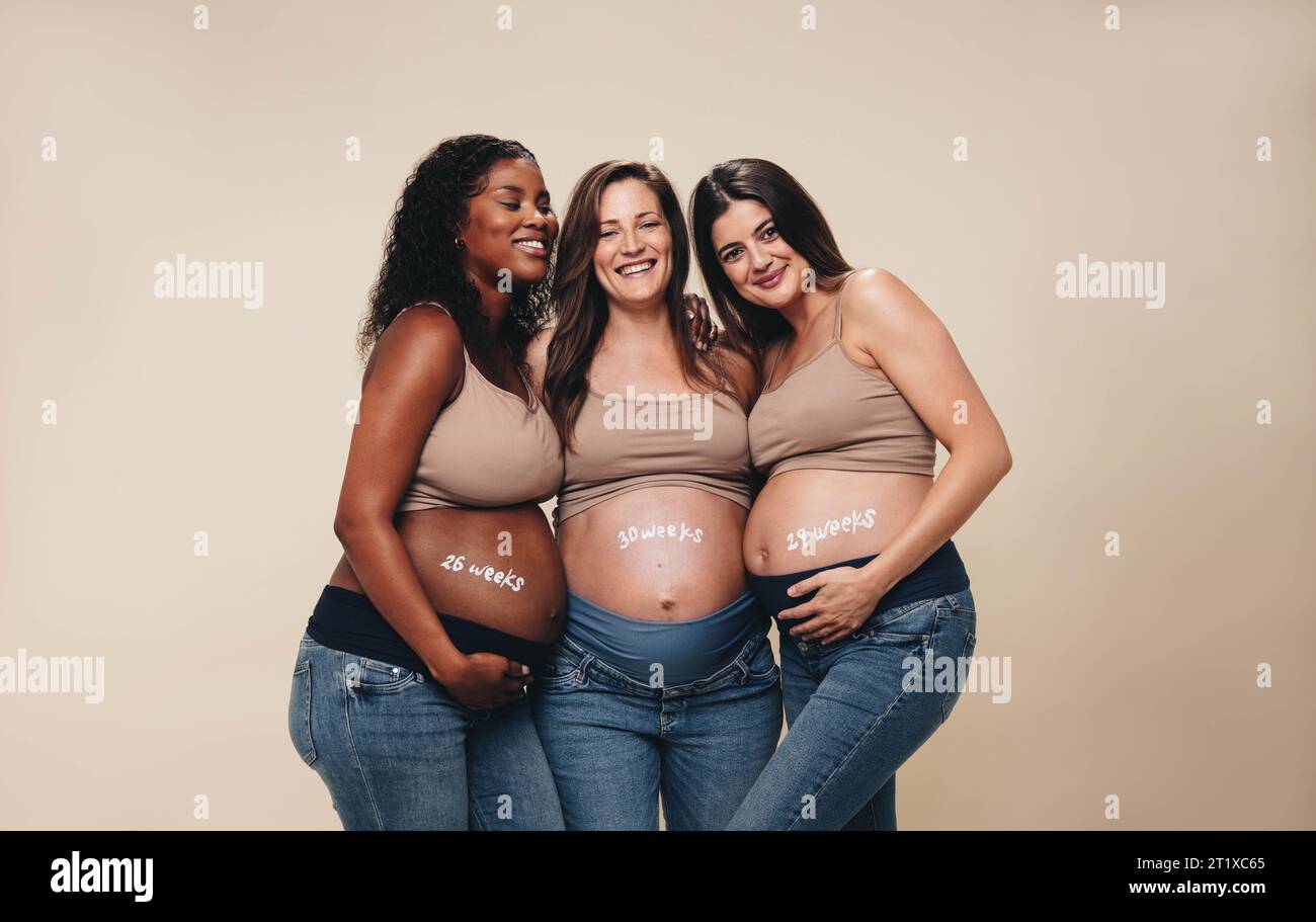 Groupe diversifié de femmes enceintes dans leur troisième trimestre debout ensemble dans un studio. Ils sourient et mettent en valeur leurs bosses de bébé. Porter des jeans et su Banque D'Images