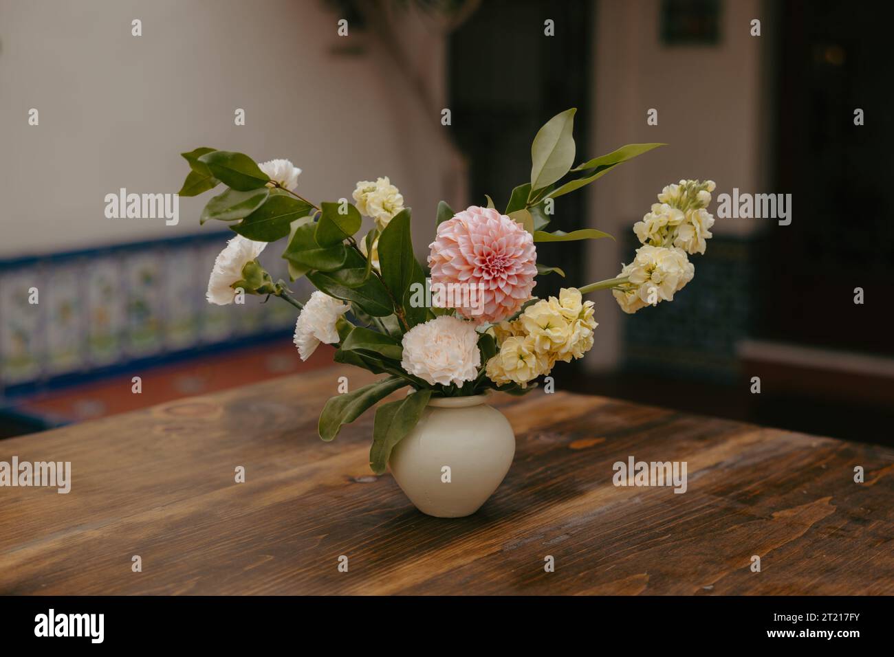 Une nature morte avec un vase en verre coloré avec un bouquet vibrant de fleurs assorties placé sur une table en bois devant un mur carrelé texturé Banque D'Images