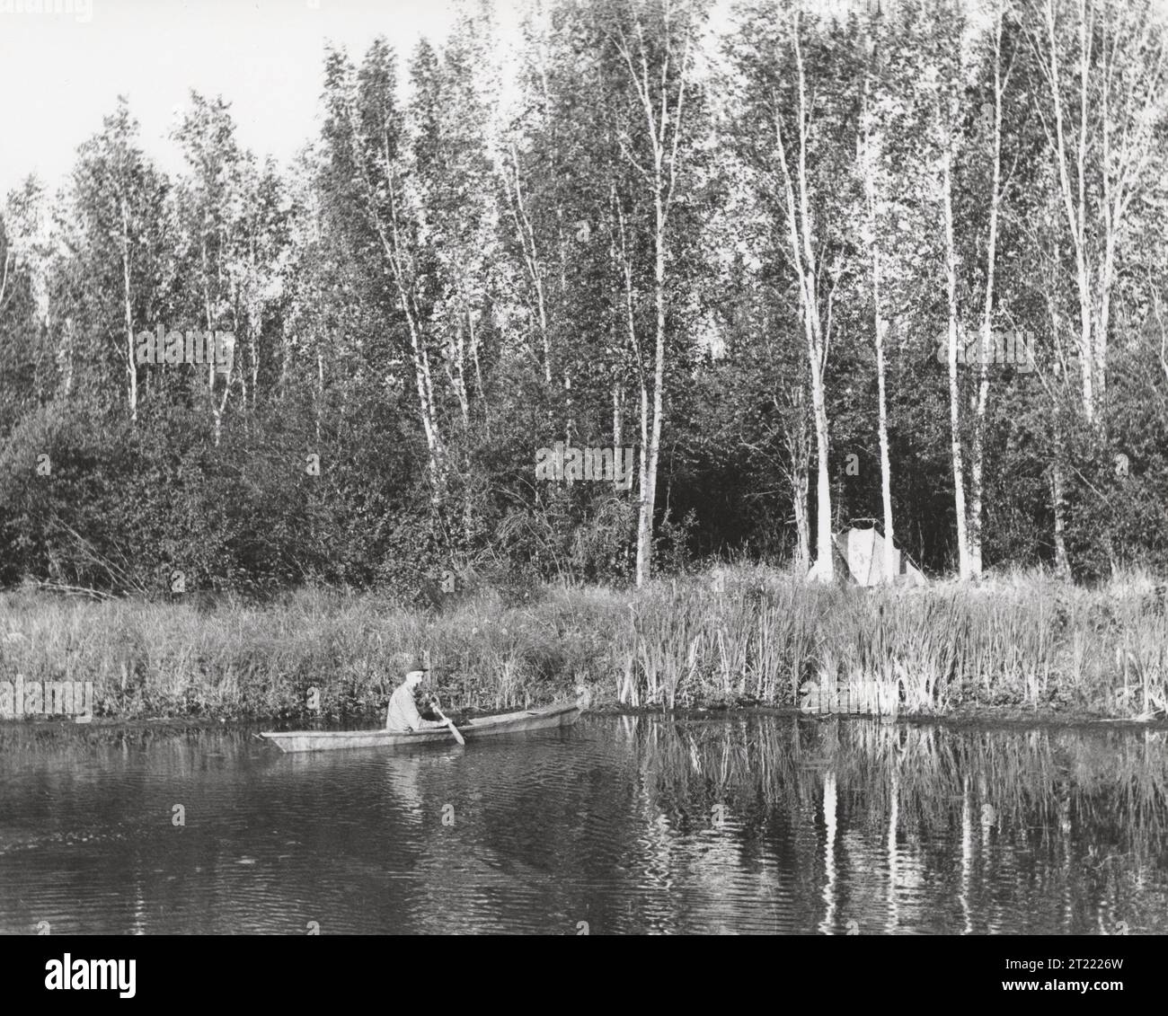 Image en noir et blanc d'un homme en canoë près d'un camp de tentes pagayant vers de hautes herbes ; NOTE : le titre original incluait 'pH-King'. Matières : Histoire ; Photographie ; Transports ; Loisirs ; Camping; canotage. Localisation : Alaska. . 1998 - 2011. Banque D'Images