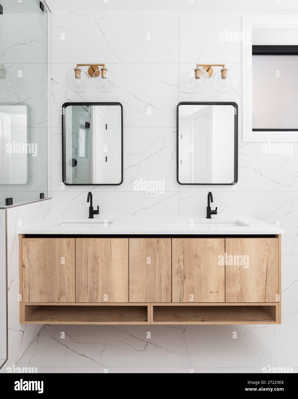 Un détail de salle de bains avec une armoire flottante en bois, un mur et un sol carrelés de marbre blanc, et un luminaire en or monté au-dessus du miroir de cadre noir. Banque D'Images